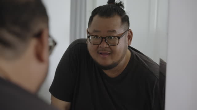 Medium shot of a large build Asian man with glasses, checking himself out in the mirror while doing positive self talk