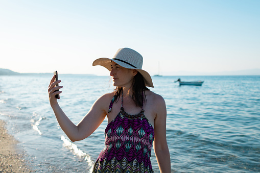 Woman with a hat is enjoying her weekend at the beach at sunset. 
She has a video call or taking selfie.