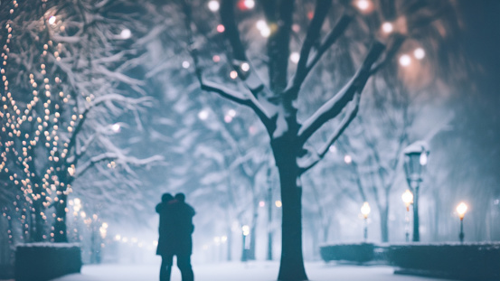 Silhouette of two lovers hugging in an atmospheric winter park with Christmas lights on trees in the evening. Blurred defocused Christmas image.