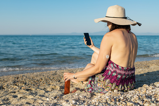 Woman with a hat is enjoying her weekend at the beach at sunset. \nShe has a video call or taking selfie. Woman has diabetes and she is wearing small sensor on the back of upper arm for controlling blood sugar.