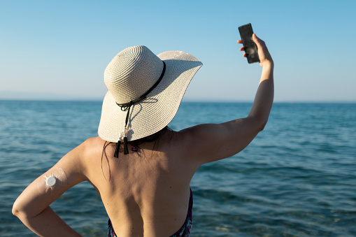 Woman with a hat is enjoying her weekend at the beach at sunset. 
She has a video call or taking selfie. Woman has diabetes and she is wearing small sensor on the back of upper arm for controlling blood sugar.