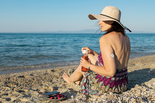 Woman with a hat is enjoying her weekend at the beach at sunset. 
She is sitting on the beach. Woman has diabetes and she is wearing small sensor on the back of upper arm for controlling blood sugar.