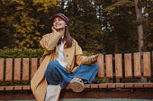 Cheerful woman enjoying music and smiling on park bench in autumn