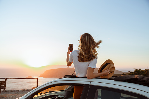 A photo of an unrecognizable woman looking at the sea and mountains from the opened roof of her car. She is taking photos of a beautiful sunset.