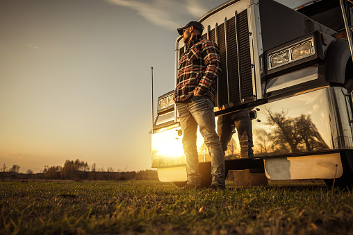 Heavy Duty Transportation Concept with Trucker in Front of His Semi Truck During Sunset. Industrial Theme.