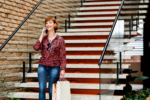 Cheerful mature woman walking down the steps carrying shopping bags talking on mobile phone and smiling