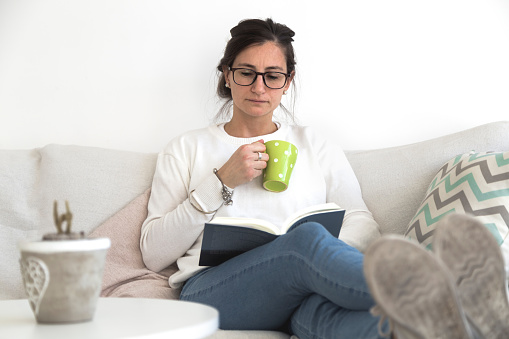Young woman sitting on sofa and reading book. Woman alone at her home reading a book with a cup in hand. She is comfortably sitting in the armchair.