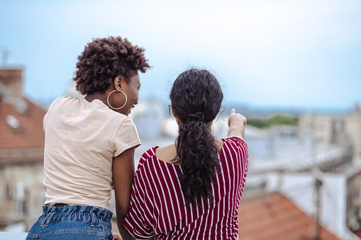 A young African American woman is standing next to a young Latin American woman who's pointing at the cityscape. They're standing with their backs turned to the camera.