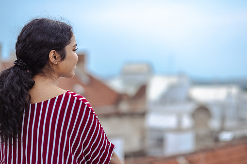 A young Latin American woman is standing with her back turned to the camera and looking at the cityscape off of a building rooftop.