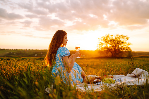 Happy woman in beautiful blue dress on picnic enjoying sunset light. Cheerful young female tourist on picnic having fun on green meadow. Concept of people, recreation, nature. Active lifestyle.