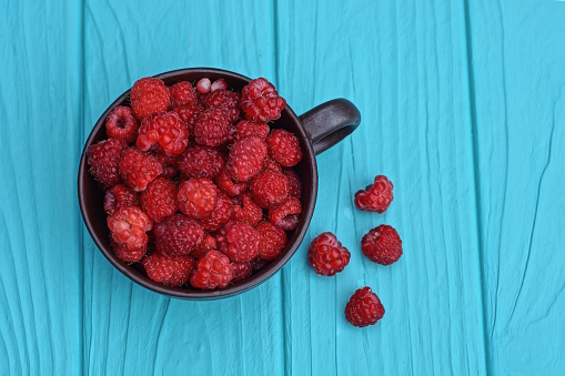 many red ripe raspberries in a brown cup on a blue wooden table