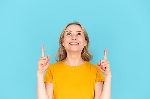 Happy woman pointing with forefingers up, on copy space for advertising and sale information. Smiling female isolated on blue background with empty place for shopping discount
