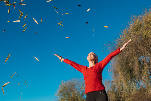 Woman beating cancer is with her arms open throwing dry leaves under the blue sky. She wears a cancer scarf and a red sweater.