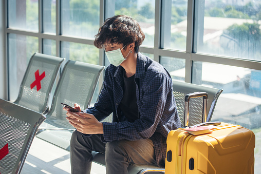 Asian young man traveller new normal wearing face mask sitting Social distancing holding smartphone in Airplane lounge. New normal Male travel pandemic passenger by plane. Coronavirus tourism trip