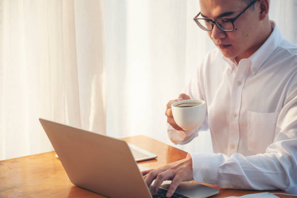 istock Asian Man drinking coffee cup using laptop at home office. Handsome man wear eyeglass holding smart tablet look website. Freelance Men using laptop home office technology. Businessman online meeting 1561866513