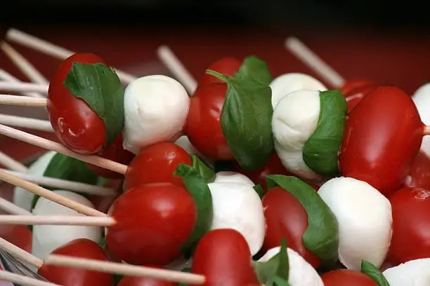 A macro photography of an italian snack composed of cherry tomatoes, tiny mozzarella balls and basil on wooden skewers.