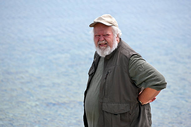 A senior gray haired man, with hand on his hip, looking to camera right, and a blurred water background.