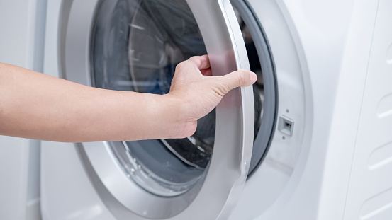 Male hand opening automatic washing machine door in laundry room.