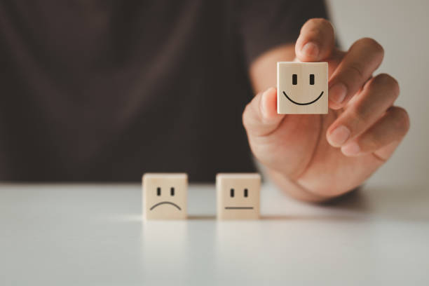 istock Customer satisfaction concept. Customer's hand lifts smiling face on blurred sad face icon on wooden cube block, Service rating, customer service best excellent business rating experience survey. 1562102571