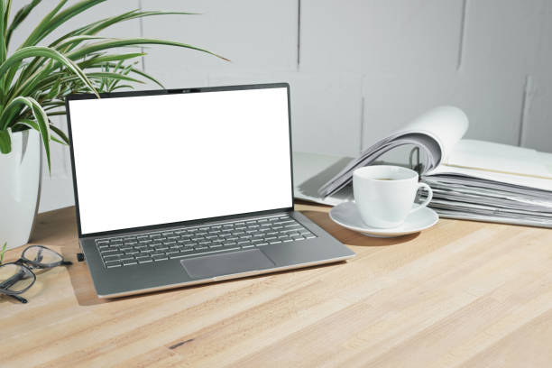 istock Mock up on an open laptop with a blank white screen, coffee cup and ring binder on a wooden office desk against a white painted wall, business concept, copy space 1562366161