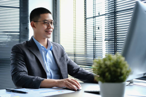 элегантный азиатский бизнесмен в очках смотрит на монитор компьютера, проводя онлайн-анализ рынка данных на современном рабочем месте. - проводя поиск стоковые фото и изображения