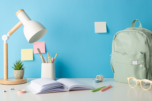 Academic ambiance captured in side view shot of well-organized desk with school essentials, eyewear, lamp, backpack, flowerpot. Blue wall backdrop with sticky notes leaves room for text or promotions