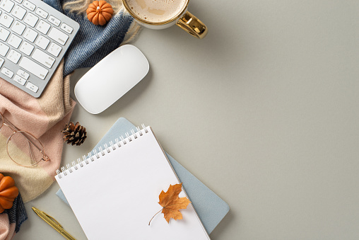 Working from home in autumn concept. Top view photo of notepad with gilded pen and glasses, keyboard and mouse, hot coffee, patchy scarf and maple leaves on grey isolated background with copyspace