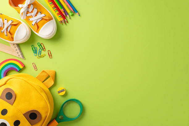 istock Get set for primary school. Top view of cute backpack shaped like a playful bear, pins, ruler, clips, magnifier, color pencils, plasticine, sneakers on soft green backdrop with room for text or ad 1562399859