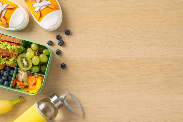 istock School lunchbox arrangement emphasizing wellness. Top view of plastic lunch container featuring assortment of fresh eating, water bottle, sneakers, placed on wooden desk, space for text or advert 1562405681