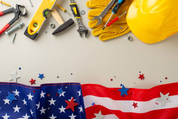 istock Labor Day with construction workers: top-view photo, displaying a flag, helmet, gloves, and building instruments on a light grey isolated background. Perfect for Labor Day marketing or text placement 1562411560
