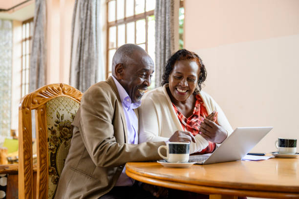 Black couple drinking coffee and using laptop together Happy man and woman in early 60s seated side by side at dining table and smiling as they review home finances and retirement savings. kenyan man stock pictures, royalty-free photos & images