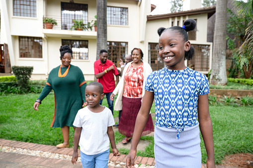 Focus on foreground 6 and 9 year old children approaching camera, followed by adults in 20s, 30s, and 60s, Nairobi two-story house in background.