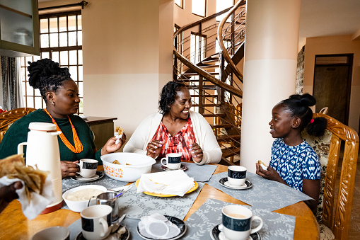 Waist-up view of 9 year old girl, late 20s mother, and early 60s grandmother sitting together at dining table in Nairobi home, talking and smiling.