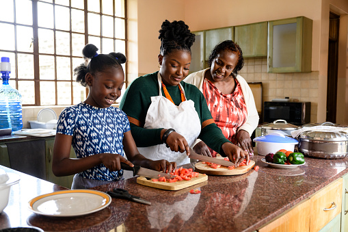 Waist-up view of 9 year old girl standing at kitchen counter in Nairobi home and slicing tomatoes with woman in late 20s, smiling woman in early 60s watching.
