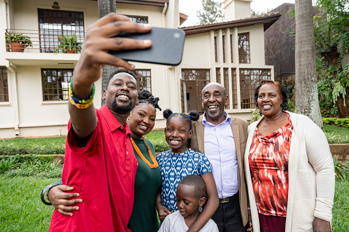 Front view of mid adult man holding smart phone, joined by three generations of adults and children, capturing memory with two-story house in background.