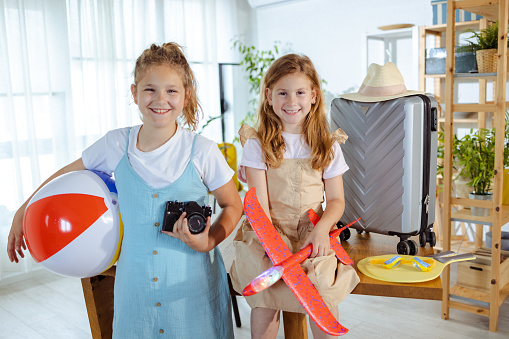 Two young Caucasian girls are cheerfully looking at the camera, while holding the things they're bringing on vacation.