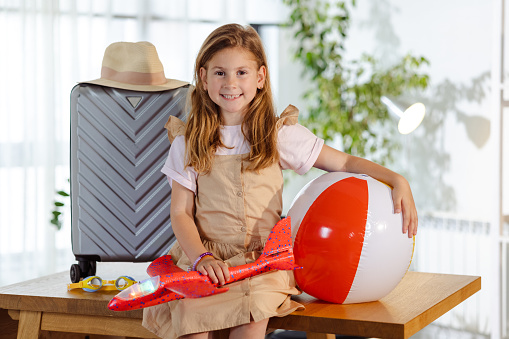 A young Caucasian girl is cheerfully looking at the camera, while holding a plane toy and a beach ball. There's a suitcase behind her.