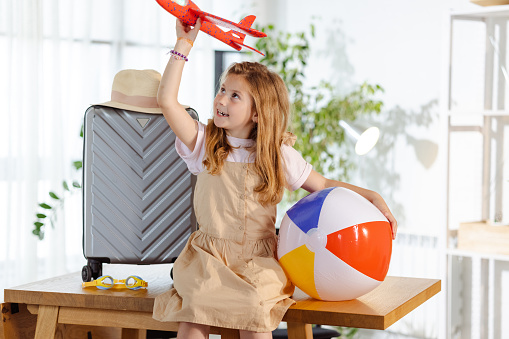 A young Caucasian girl is flying a plane toy over her head and holing a beach ball in the other hand. There's a suitcase behind her.