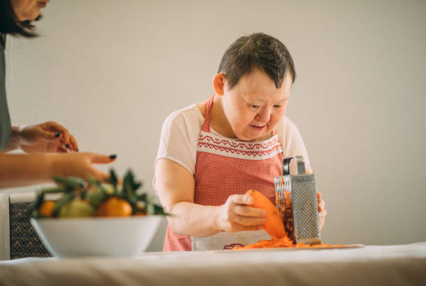 elderly woman with Down syndrome gratefully learns to grate carrots with the guidance of a teacher stock photo