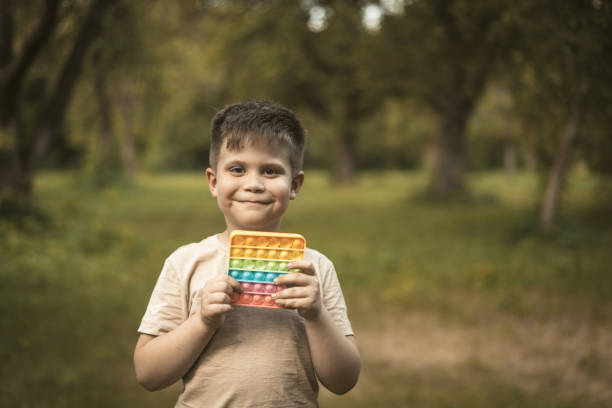 istock Smiling Boy With Rainbow Pop It Walks in the Park. Nice Kid Plays Pop It Outdoor. 1562637097