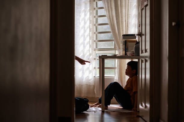 A little Asian boy with a sad and stress expression hides in the shadows under a table at home, while being scolded by his mother