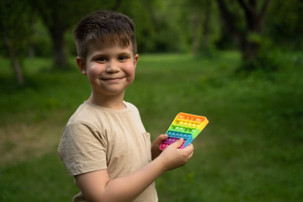 istock Pink-Cheeked Boy with Nice Dimples Clicks his New Pop It. Smiling 7 years guy having a walk in the park. Park Background. 1562643910