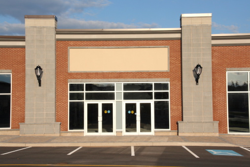 Blank generic brick storefront in modern strip mall with vertical columns and glass doors, suitable for retail concept mock-ups.  Includes drive up parking markings and disabled parking spot.  Blue sky, sunny day, lamp lights.