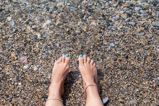 Serene photo featuring young woman's feet playfully dancing in the lapping waves of the sea, adorned with delicate anklets, evoking a sense of relaxation and summer joy.