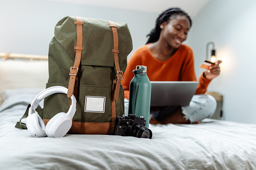 A young African American woman is shopping on her laptop in the back, while her backpack is in the forefront. Next to it are wireless headphones, a camera and a water bottle.