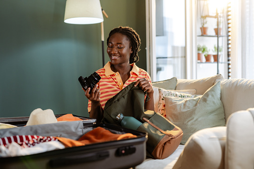 A young African American woman is holding a camera, while packing her backpack for a trip. There's an open suitcase in front of her.
