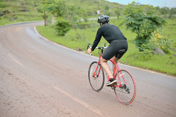Rear view of a determined cyclist gracefully pedaling along a serene country road. As the rider ventures through the picturesque countryside, the image showcases their athletic silhouette, accentuating their passion for cycling and love for outdoor adventures. The idyllic rural setting, with lush fields, creates a sense of peace and tranquility, highlighting the joy of cycling amidst nature's embrace.
