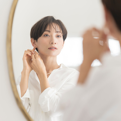 An Asian woman is getting dressed. She is looking at a mirror and  wearing accessories. earrings.