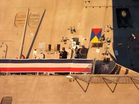 An alert armed guard patrols the stern flight deck the USS Canberra (LCS 30) docked at Garden Island in Sydney Harbour.  USS Canberra is an Independence Class combat ship of the US Navy.  She is in port for her commissioning ceremony on 22 July 2023.  She is adorned with red, white and blue bunting.  A sign behind the guard reads, \
