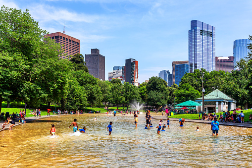 Boston, Massachusetts, USA - July 19, 2023: The Boston Common Frog Pond sits at the heart of Boston Common. Managed by The Skating Club of Boston in partnership with the City of Boston, Frog Pond is home to a winter ice skating rink and learn-to-skate school, a reflecting pool in the spring and fall, and a summer spray pool. Downtown Boston skyscrapers in the background.